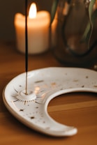 A lit candle is placed on a wooden surface next to a decorative incense holder shaped like a crescent moon. The incense stick is vertical, emitting a thin trail of smoke. In the background, there is a glass jar with plant leaves.