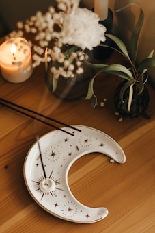 A wooden table decorated with candles, a white moon-shaped dish with golden celestial designs, and a couple of sticks of incense. A white flower and some green leaves are also present, creating a serene and aesthetic arrangement.