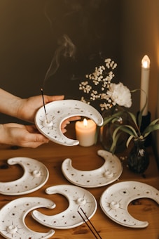 A set of crescent-shaped ceramic incense holders with star and moon designs are placed on a wooden table. One is held by a hand, with an incense stick burning and smoke rising. In the background, a lit candle, small white flowers, and a tall candle contribute to the warm ambiance.
