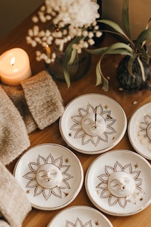 An arrangement of ceramic plates with sun designs featuring serene faces, positioned on a wooden surface. A lit candle, a knitted fabric, and vases containing greenery and a bouquet of white flowers add to the cozy ambiance.