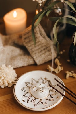 A serene setup featuring a lit candle in the background, a decorative ceramic plate with a sun face design, and incense sticks resting on the plate. The smoke from the incense adds a calming atmosphere. Surrounding elements include a cozy blanket, some green leaves, a white chrysanthemum flower, and a few dried flower petals.