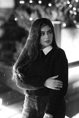 A monochrome portrait of a woman with long hair, standing with one arm crossed over the other. The background has soft bokeh lights, creating a blurred effect that highlights the subject. She wears a dark sweater and a white collared shirt beneath, with a contemplative expression.