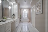 A spacious and elegant bathroom features a long white vanity with a large mirror and modern lighting. The room has a bathtub situated under a window with sheer curtains, a decorative plant, and a chandelier overhead. Neutral tones and clean lines create a sophisticated and serene atmosphere.