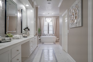A spacious and elegant bathroom features a long white vanity with a large mirror and modern lighting. The room has a bathtub situated under a window with sheer curtains, a decorative plant, and a chandelier overhead. Neutral tones and clean lines create a sophisticated and serene atmosphere.