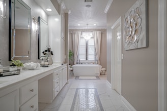 A spacious and elegant bathroom features a long white vanity with a large mirror and modern lighting. The room has a bathtub situated under a window with sheer curtains, a decorative plant, and a chandelier overhead. Neutral tones and clean lines create a sophisticated and serene atmosphere.