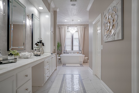 A spacious and elegant bathroom features a long white vanity with a large mirror and modern lighting. The room has a bathtub situated under a window with sheer curtains, a decorative plant, and a chandelier overhead. Neutral tones and clean lines create a sophisticated and serene atmosphere.