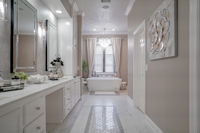 A spacious and elegant bathroom features a long white vanity with a large mirror and modern lighting. The room has a bathtub situated under a window with sheer curtains, a decorative plant, and a chandelier overhead. Neutral tones and clean lines create a sophisticated and serene atmosphere.