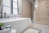 A modern, elegant bathroom featuring a large white bathtub placed near a window with blinds. Next to the bathtub, there are two decorative candle holders and a soap dish. The floor is tiled with a light, marble-like pattern, matching the shower enclosure to the right. A glass chandelier hangs above, adding a touch of luxury. A simple towel is draped over the tub, and there is a plant and a candle on a small table in the foreground.