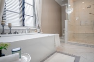 A modern, elegant bathroom featuring a large white bathtub placed near a window with blinds. Next to the bathtub, there are two decorative candle holders and a soap dish. The floor is tiled with a light, marble-like pattern, matching the shower enclosure to the right. A glass chandelier hangs above, adding a touch of luxury. A simple towel is draped over the tub, and there is a plant and a candle on a small table in the foreground.