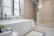 A modern, elegant bathroom featuring a large white bathtub placed near a window with blinds. Next to the bathtub, there are two decorative candle holders and a soap dish. The floor is tiled with a light, marble-like pattern, matching the shower enclosure to the right. A glass chandelier hangs above, adding a touch of luxury. A simple towel is draped over the tub, and there is a plant and a candle on a small table in the foreground.