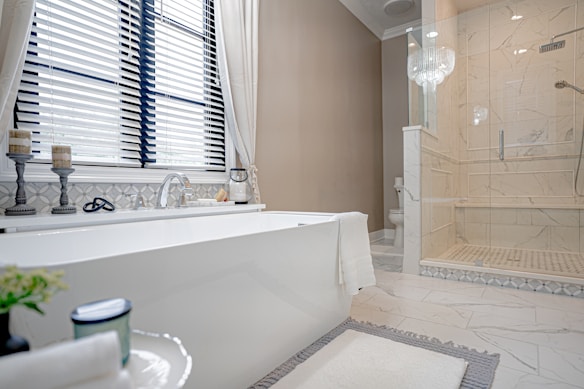 A modern, elegant bathroom featuring a large white bathtub placed near a window with blinds. Next to the bathtub, there are two decorative candle holders and a soap dish. The floor is tiled with a light, marble-like pattern, matching the shower enclosure to the right. A glass chandelier hangs above, adding a touch of luxury. A simple towel is draped over the tub, and there is a plant and a candle on a small table in the foreground.