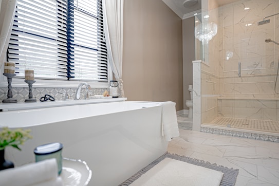 A modern, elegant bathroom featuring a large white bathtub placed near a window with blinds. Next to the bathtub, there are two decorative candle holders and a soap dish. The floor is tiled with a light, marble-like pattern, matching the shower enclosure to the right. A glass chandelier hangs above, adding a touch of luxury. A simple towel is draped over the tub, and there is a plant and a candle on a small table in the foreground.