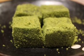 Four green, matcha-dusted cubes are arranged closely together on a dark plate. The texture appears soft and crumbly, while the color is a vibrant green.