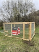 A wooden animal enclosure is set on a grassy area, featuring a vibrant red mini barn-style structure inside. The enclosure is reinforced with mesh wire and sturdy wooden beams, and is surrounded by leafless trees in the background, suggesting early spring or late fall.