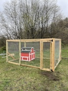 A wooden animal enclosure is set on a grassy area, featuring a vibrant red mini barn-style structure inside. The enclosure is reinforced with mesh wire and sturdy wooden beams, and is surrounded by leafless trees in the background, suggesting early spring or late fall.