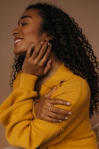 A person with long, curly hair is wearing a yellow sweater, gently touching their neck and shoulder. They are adorned with earrings and rings, and have a calm and joyful expression on their face.