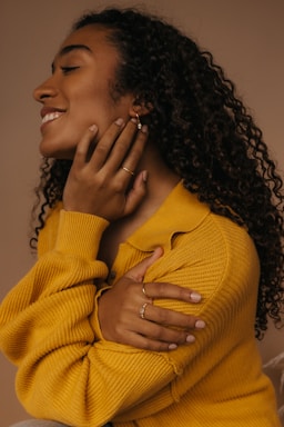 A person with long, curly hair is wearing a yellow sweater, gently touching their neck and shoulder. They are adorned with earrings and rings, and have a calm and joyful expression on their face.