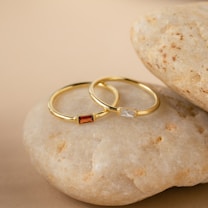 Two gold rings with rectangular gemstones, one red and one clear, placed on a smooth beige stone surface. The background complements the stones with a matching sandy hue, creating a soft and elegant atmosphere.