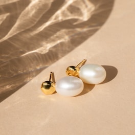 A pair of elegant pearl earrings with gold accents rests on a smooth beige surface. Soft shadows and light reflections create a subtle, sophisticated pattern across the background.