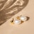 A pair of elegant pearl earrings with gold accents rests on a smooth beige surface. Soft shadows and light reflections create a subtle, sophisticated pattern across the background.