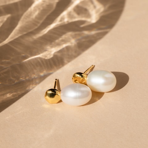 A pair of elegant pearl earrings with gold accents rests on a smooth beige surface. Soft shadows and light reflections create a subtle, sophisticated pattern across the background.