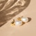 A pair of elegant pearl earrings with gold accents rests on a smooth beige surface. Soft shadows and light reflections create a subtle, sophisticated pattern across the background.