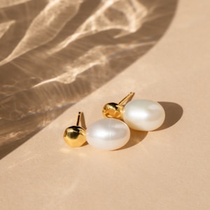 A pair of elegant pearl earrings with gold accents rests on a smooth beige surface. Soft shadows and light reflections create a subtle, sophisticated pattern across the background.