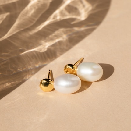 A pair of elegant pearl earrings with gold accents rests on a smooth beige surface. Soft shadows and light reflections create a subtle, sophisticated pattern across the background.