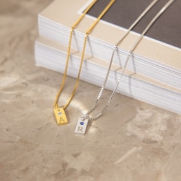 Two necklaces with rectangular pendants lie on a marble surface. One pendant is gold with the letter 'A' and features a small jewel. The other is silver with the letter 'R' and a blue gem. The chains are thin and metallic, extending diagonally across the image. Behind them, a stack of closed books with a beige and white cover is visible.