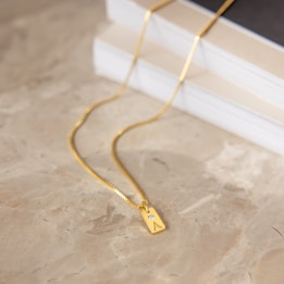 A gold necklace with a rectangular pendant lying on a marble surface. The pendant features the letter 'A' engraved along with a small diamond and a book in the background.