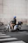 A person wearing a blue helmet is sitting on a white electric scooter parked near a curb. The scooter has a storage box on the back and is positioned next to a pedestrian crosswalk. The backdrop is a large stone wall, creating a contrast to the urban street setting.