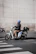 A person wearing a blue helmet is sitting on a white electric scooter parked near a curb. The scooter has a storage box on the back and is positioned next to a pedestrian crosswalk. The backdrop is a large stone wall, creating a contrast to the urban street setting.