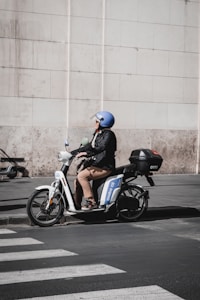 A person wearing a blue helmet is sitting on a white electric scooter parked near a curb. The scooter has a storage box on the back and is positioned next to a pedestrian crosswalk. The backdrop is a large stone wall, creating a contrast to the urban street setting.