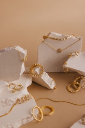 A collection of gold jewelry, including chains, rings, and bracelets, is artfully arranged on pieces of rough white stone. The jewelry features various designs and some pieces are adorned with small stones or beads. The backdrop is a neutral, warm beige color that complements the gold tones.