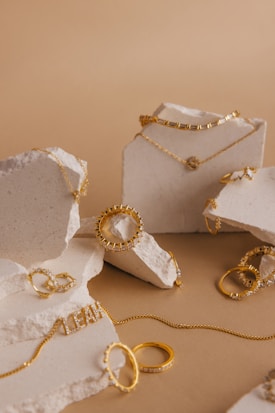 A collection of gold jewelry, including chains, rings, and bracelets, is artfully arranged on pieces of rough white stone. The jewelry features various designs and some pieces are adorned with small stones or beads. The backdrop is a neutral, warm beige color that complements the gold tones.