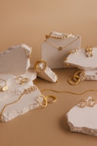 A collection of gold jewelry pieces including necklaces, rings, and earrings is artfully arranged on pieces of textured stone against a light brown background. The jewelry features delicate chain links, engravings, and intricate detailing.