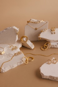A collection of gold jewelry pieces including necklaces, rings, and earrings is artfully arranged on pieces of textured stone against a light brown background. The jewelry features delicate chain links, engravings, and intricate detailing.