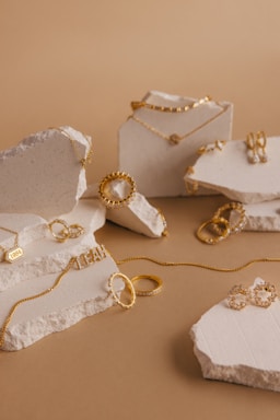 A collection of gold jewelry pieces including necklaces, rings, and earrings is artfully arranged on pieces of textured stone against a light brown background. The jewelry features delicate chain links, engravings, and intricate detailing.