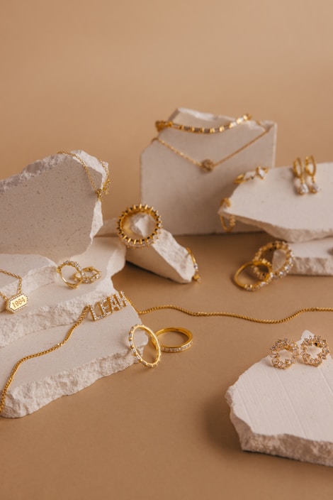 A collection of gold jewelry pieces including necklaces, rings, and earrings is artfully arranged on pieces of textured stone against a light brown background. The jewelry features delicate chain links, engravings, and intricate detailing.