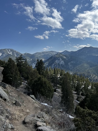 A mountainous landscape with a dirt trail winding through dense pine trees. Snow is lightly dusted on the peaks of the mountains, and the sky is partly cloudy with patches of clear blue.