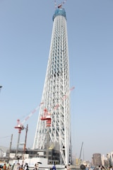A tall modern tower under construction with a lattice structure, surrounded by cranes. The lattice is white with areas covered in blue tarpaulin, and several red cranes are working on different levels. There are some buildings in the background as well as people walking around the construction area.