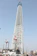 A tall modern tower under construction with a lattice structure, surrounded by cranes. The lattice is white with areas covered in blue tarpaulin, and several red cranes are working on different levels. There are some buildings in the background as well as people walking around the construction area.