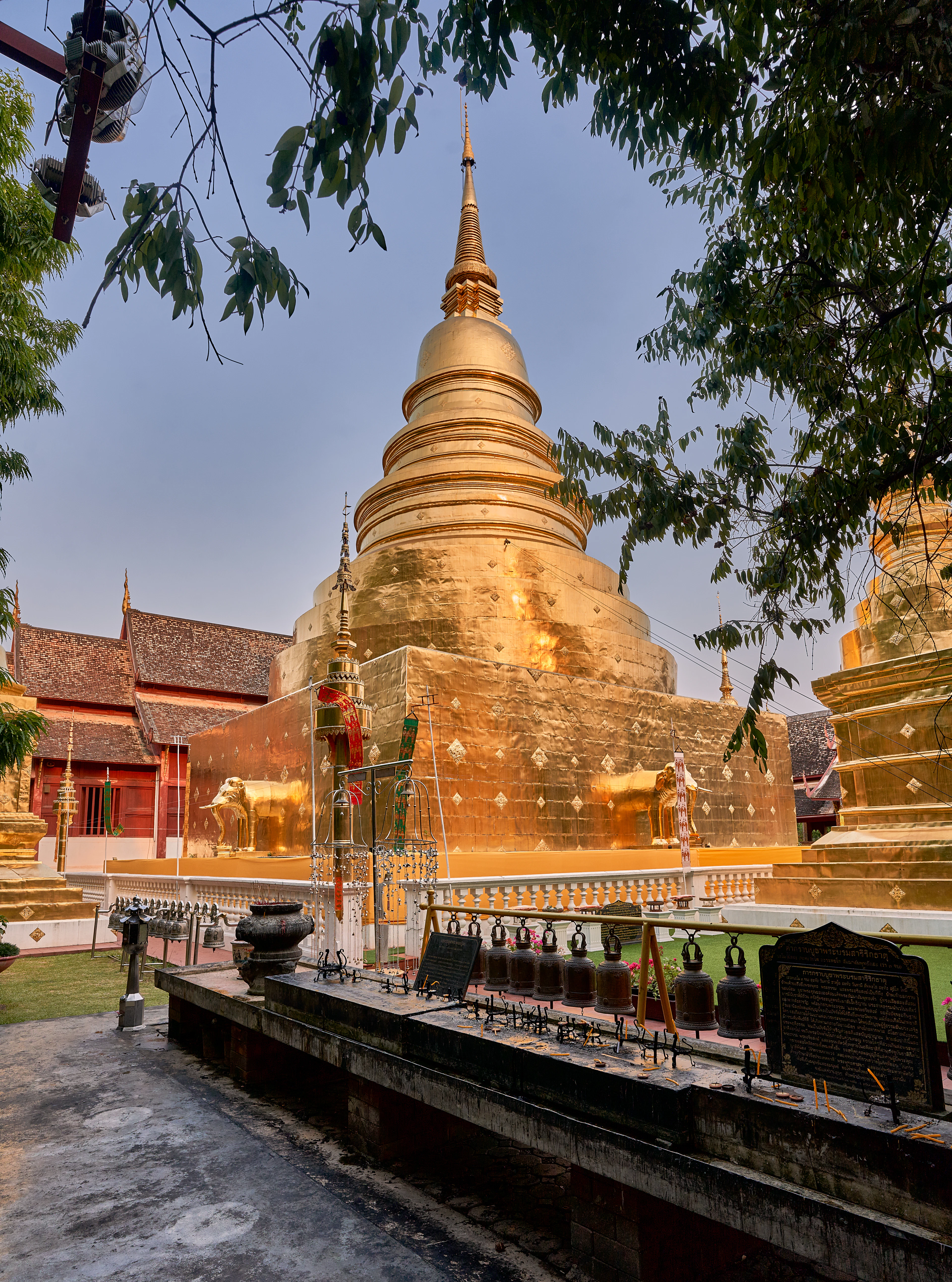 A gilded bell-shaped chedi and red-tiled Lanna temple roofs in Chiang Mai, with a long row of bronze prayer bells lining the terrace in front
