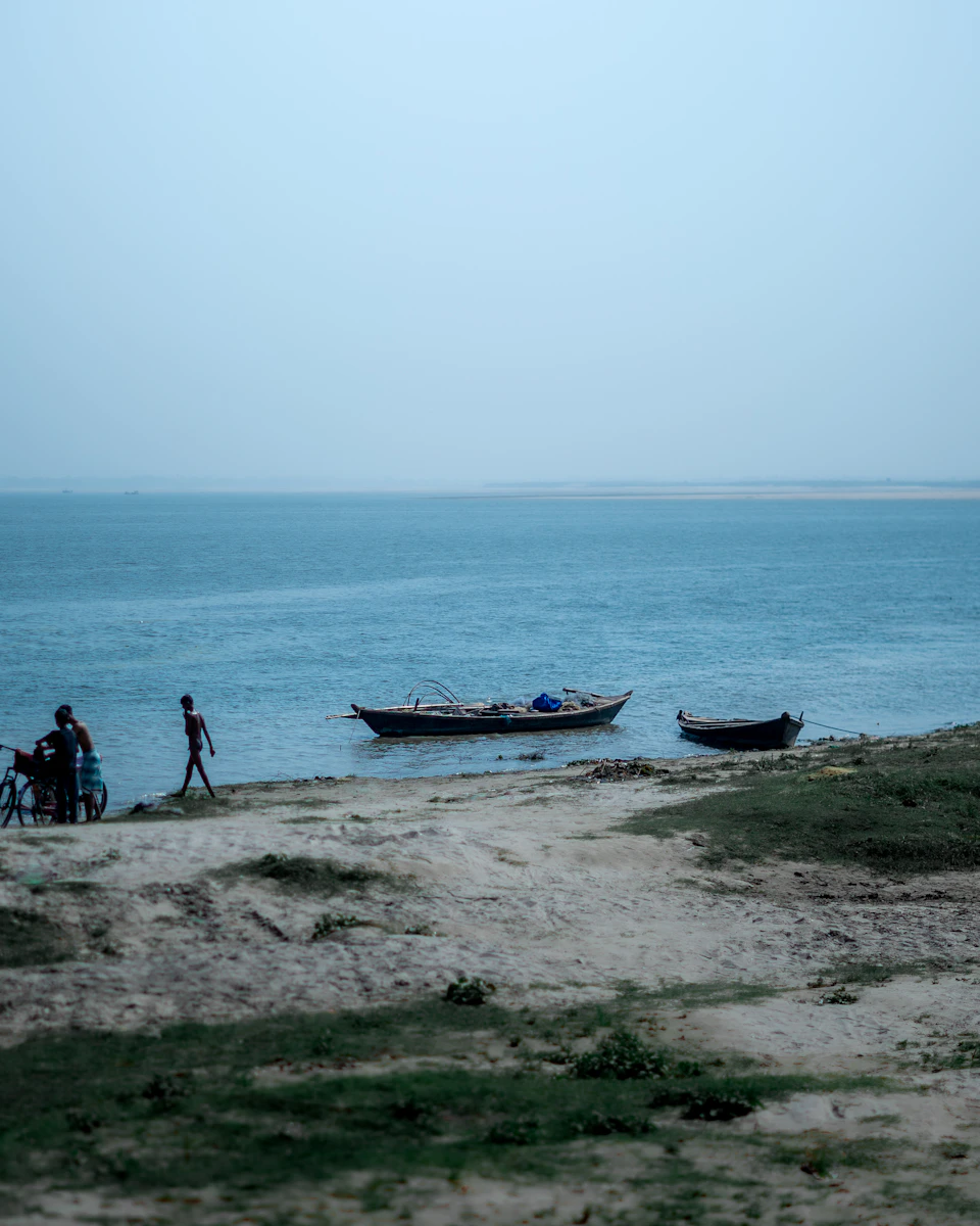 The image depicts a serene beach scene with three boats floating on the water, two small and one larger boat. The smaller boats are positioned closer to the shoreline, while the larger boat is further out in the ocean. A person can be seen walking along the sandy beach near the water's edge. In the background, there is a grassy area that extends towards the horizon where it meets the sky.