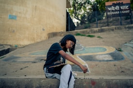 A young person is sitting on a concrete slope, focused intently on creating artwork on the ground with brush and paint. The surroundings are outdoors, possibly near a historical or culturally significant site, as indicated by a sign that reads 'Bhadaini Ghat'. Trees and a fence are visible in the background.
