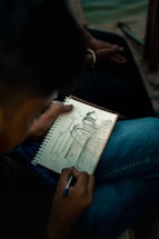A person is holding a pen and sketching a detailed architectural drawing in a spiral-bound notebook. The drawing features a building with a dome, intricate patterns, and surrounding structures. The person is seated, wearing jeans, and the setting appears to be outdoors, possibly near water.