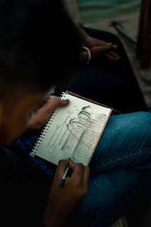 A person is holding a pen and sketching a detailed architectural drawing in a spiral-bound notebook. The drawing features a building with a dome, intricate patterns, and surrounding structures. The person is seated, wearing jeans, and the setting appears to be outdoors, possibly near water.