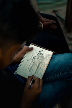 A person is holding a pen and sketching a detailed architectural drawing in a spiral-bound notebook. The drawing features a building with a dome, intricate patterns, and surrounding structures. The person is seated, wearing jeans, and the setting appears to be outdoors, possibly near water.