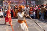 A man dressed in traditional Indian attire, featuring a white garment and an orange turban, performs with a long wooden stick while leading a parade. A lively crowd watches the display, some in festive clothing, against a backdrop of colorful banners. Confetti is scattered on the ground, adding to the celebratory atmosphere.
