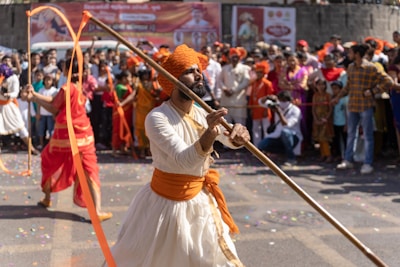 A man dressed in traditional Indian attire, featuring a white garment and an orange turban, performs with a long wooden stick while leading a parade. A lively crowd watches the display, some in festive clothing, against a backdrop of colorful banners. Confetti is scattered on the ground, adding to the celebratory atmosphere.